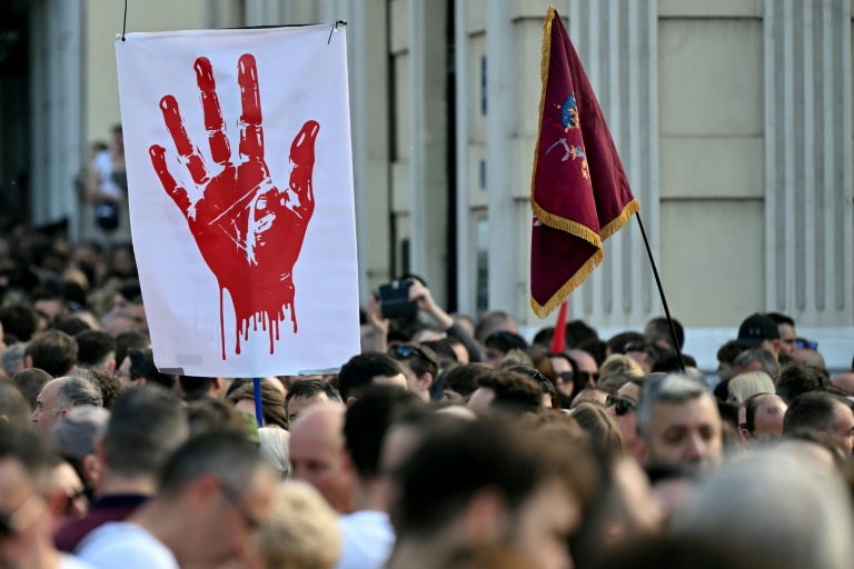 Des personnes défilent derrière une banderole ornée d’une main rouge lors d’une manifestation antigouvernementale à Belgrade, le 23 mai 2026, dénonçant la corruption et réclamant des élections anticipées après l’effondrement meurtrier de l'auvent de la gare de Novi Sad, en Serbie, en 2024