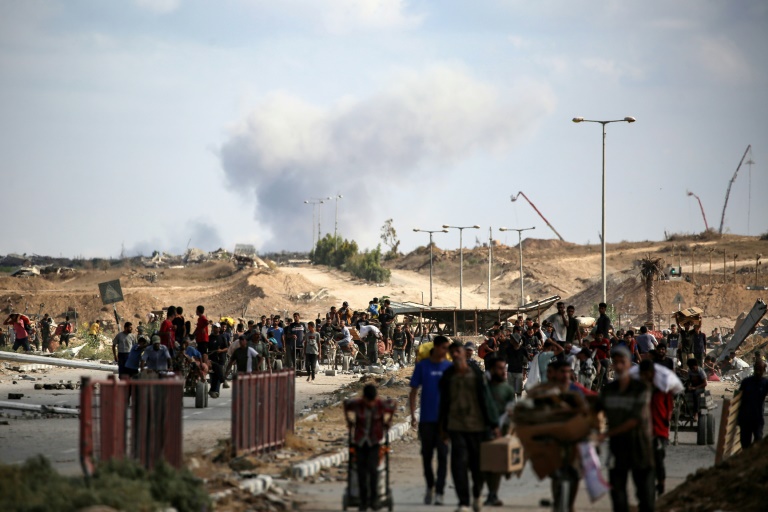 Une colonne de fumée s'élève  en 
arrière-plan après une frappe alors que des Palestiniens reviennent d'un point de distribution alimentaire dans le centre de la bande de Gaza, le 5 octobre 2025