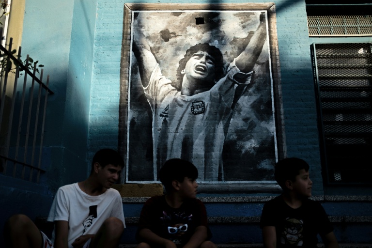 Des jeunes assis devant une fresque murale représentant la star du football décédée Diego Maradona, dans son quartier natal de Villa Fiorito, dans la province de Buenos Aires, le 9 avril 2026 en Argentine 