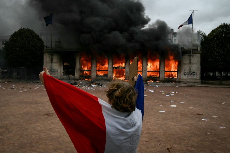 Un protestataire enveloppé dans un drapeau français devant l'antenne de l'Office du tourisme de Lyon en feu, lors d'une manifestation contre la vie chère, le 17 octobre 2026